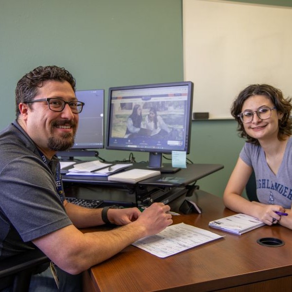 Two students stand outside a campus building labeled “Student Services.” They both carry backpacks, and one holds a booklet and papers. Trees with yellow leaves frame the scene, and the building’s large windows reflect the outdoor surroundings.