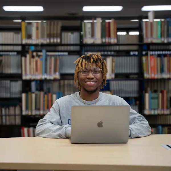 Three students sitting at an outdoor table on campus, working together with a laptop and notebooks, surrounded by trees and greenery.