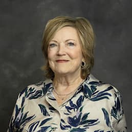 Studio portrait of a smiling older woman wearing a floral blouse against a gray background.