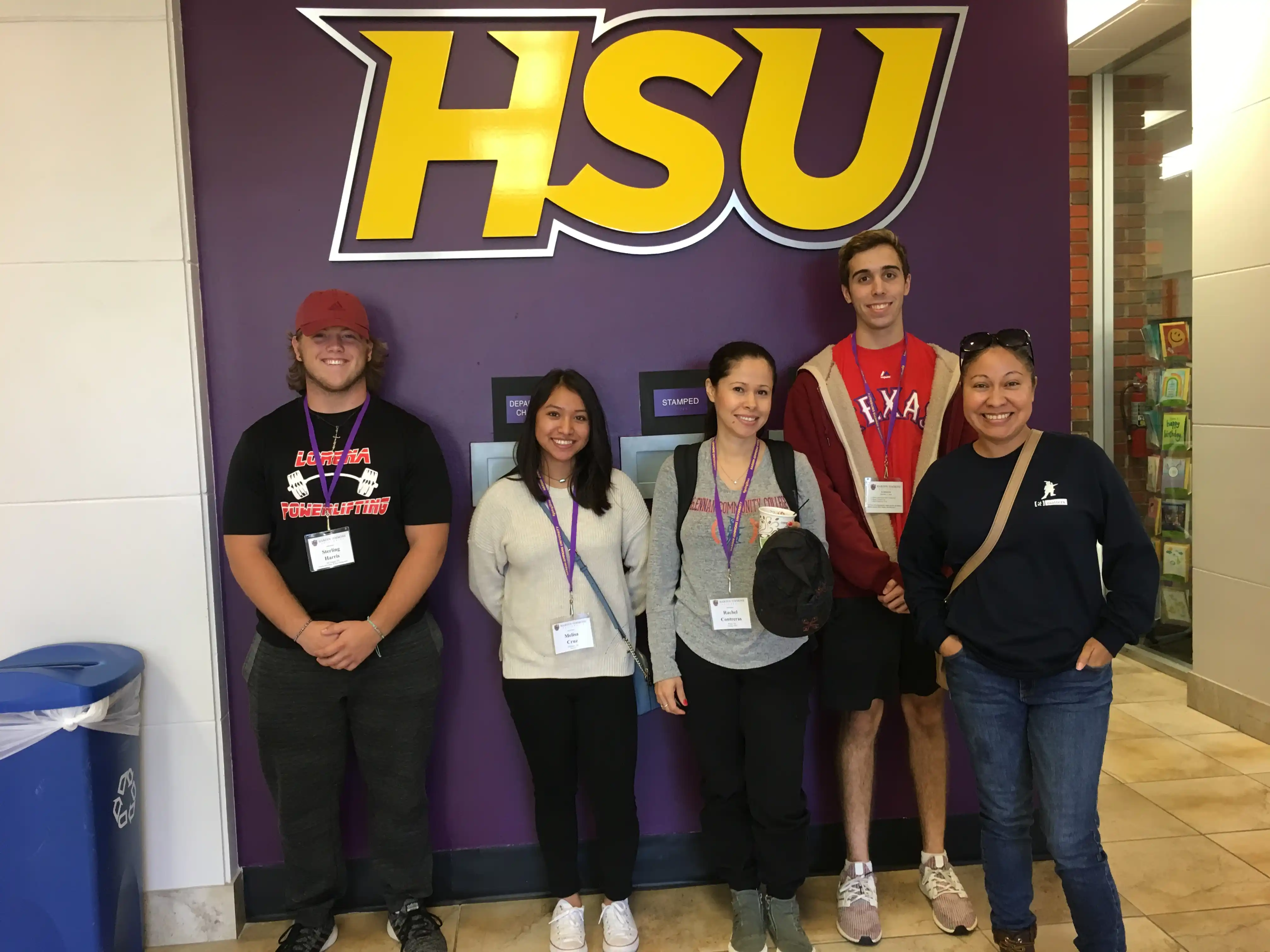 Group of five people posing in a hallway in front of a large wall sign that reads “HSU.”
