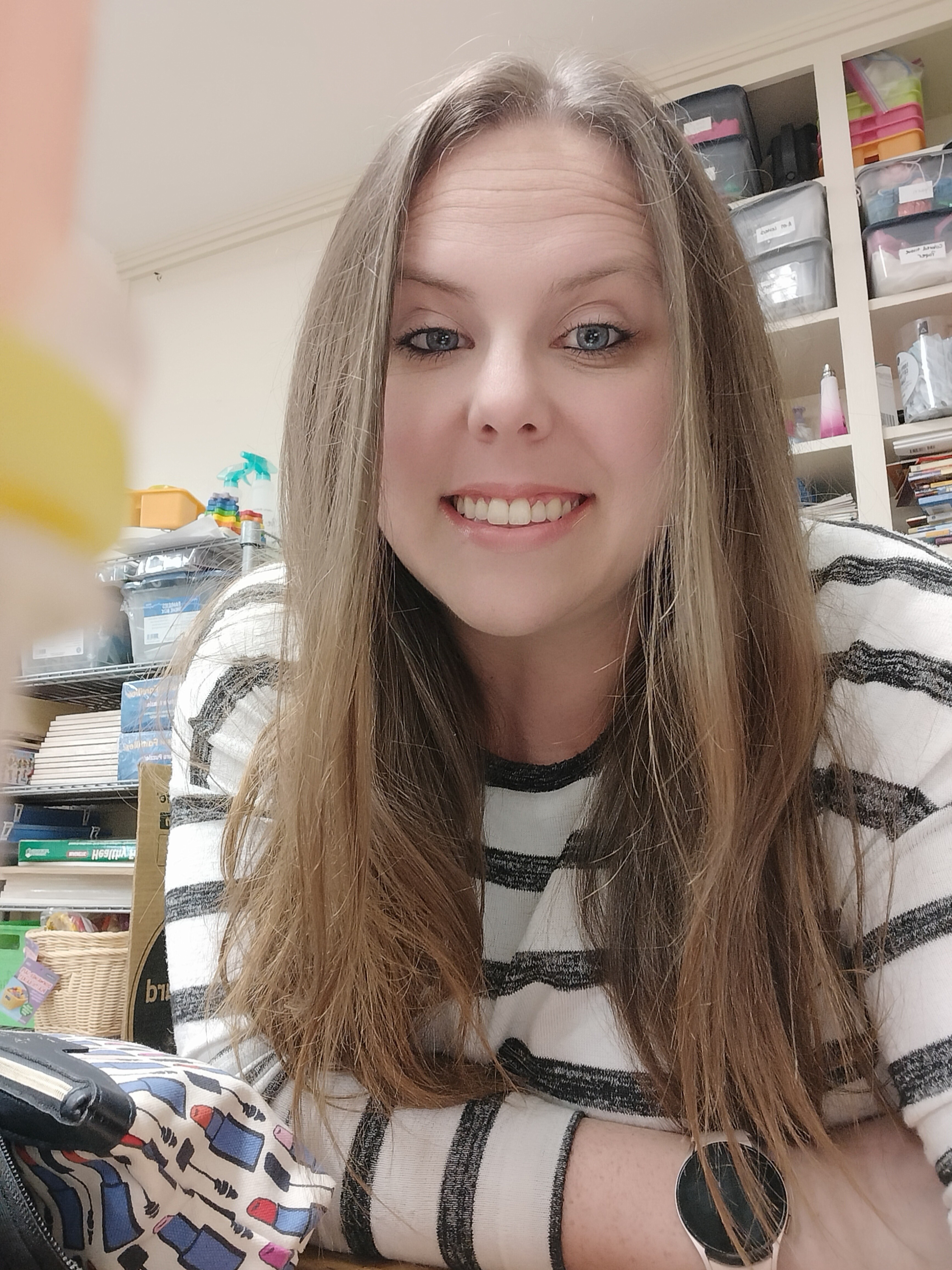 A smiling woman with long hair, wearing a striped shirt, poses for a selfie in a colorful workspace filled with art supplies.