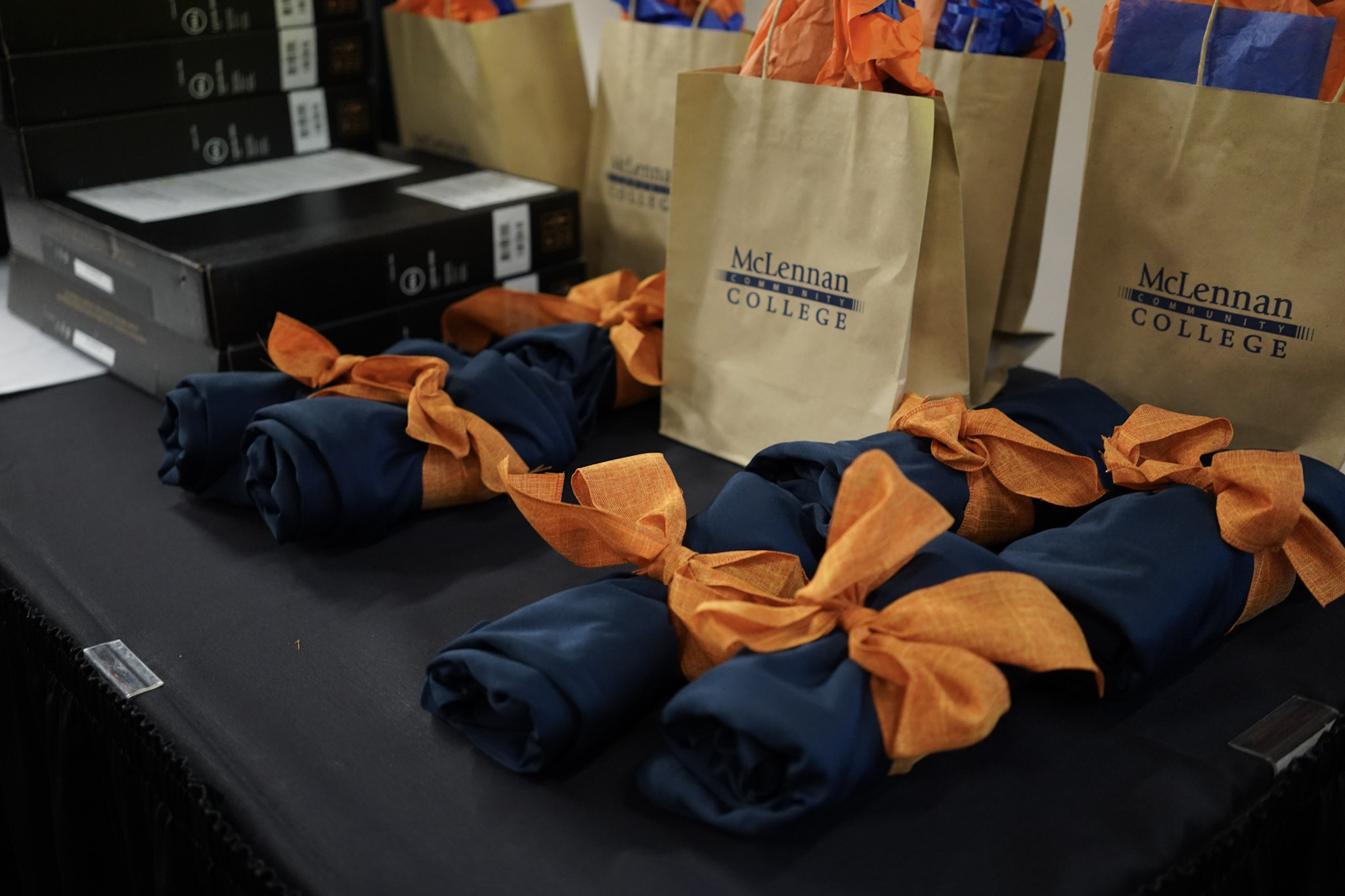 Gift bags with McLennan College branding, each tied with orange ribbons and containing rolled items, arranged on a table.