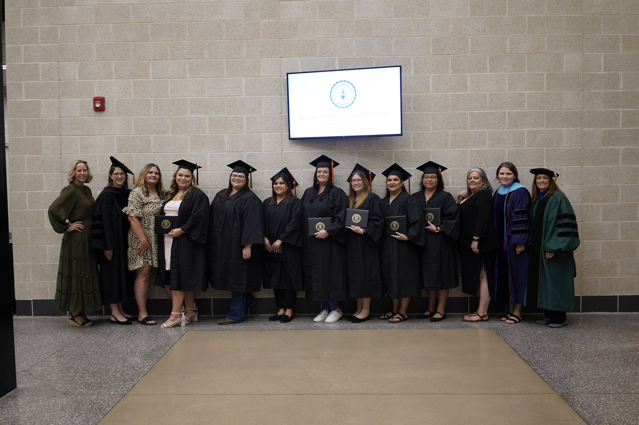 Group of graduates in caps and gowns posing with their certificates, accompanied by faculty members at a graduation ceremony.