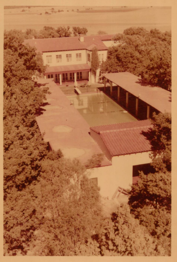 Aerial view of a mid-century building complex featuring a swimming pool and surrounding greenery.