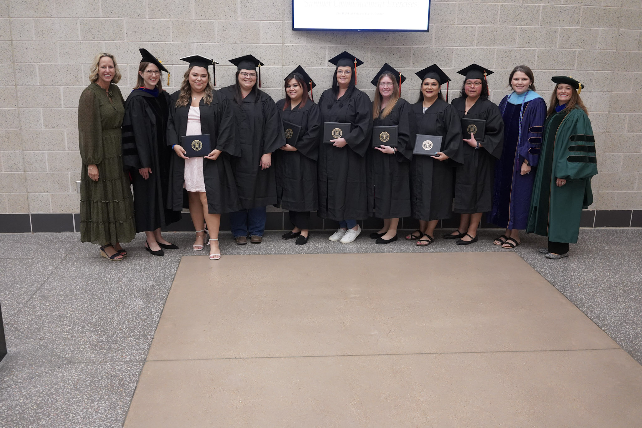 Group of graduates in cap and gown holding diplomas, posing for a photo with faculty members.