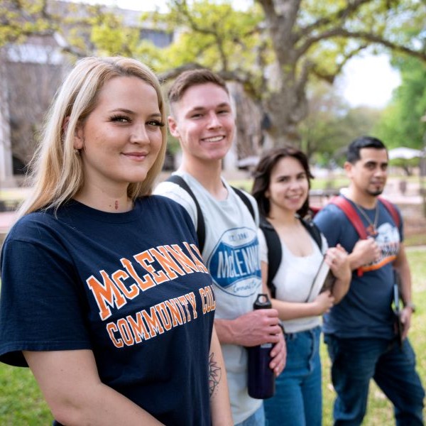 A group of four students stands outdoors on a college campus, with trees and a building in the background. The student in the foreground wears a navy McLennan Community College T-shirt. The others carry backpacks and a water bottle, appearing to be walking or gathering between classes on a sunny day.
