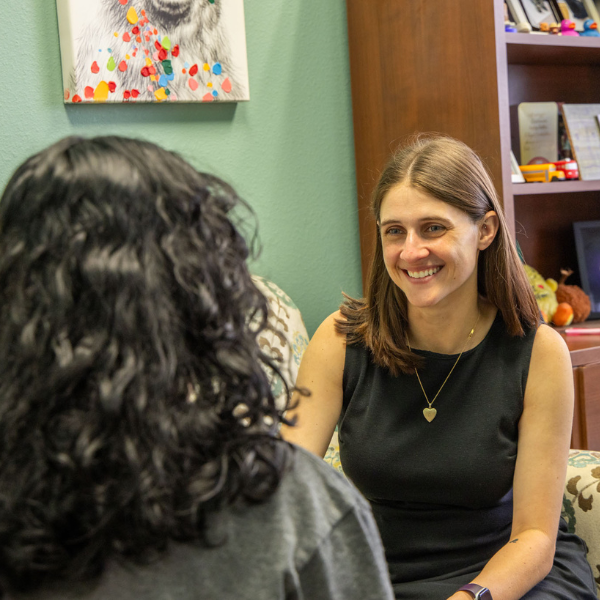 Two people seated across from each other in a small office, engaged in a one-on-one conversation. The setting includes a bookshelf with books and decor, a framed artwork on the wall, and soft, neutral furnishings, suggesting an academic advising or support meeting environment.