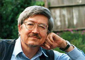 A portrait of a middle-aged man with glasses and gray hair, resting his chin on his hand in a garden setting.
