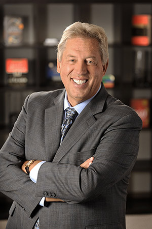 A confident businessman in a gray suit stands with arms crossed, smiling warmly against a backdrop of bookshelves.
