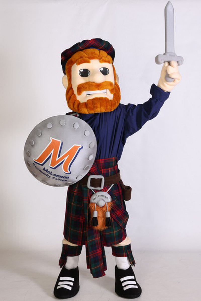 McLennan Community College's Highlander mascot, "Mac," stands proudly in a Scottish kilt and tam, holding a silver sword in one hand and a round shield featuring the MCC logo in the other. He has a large orange beard and an expressive cartoon-style face.