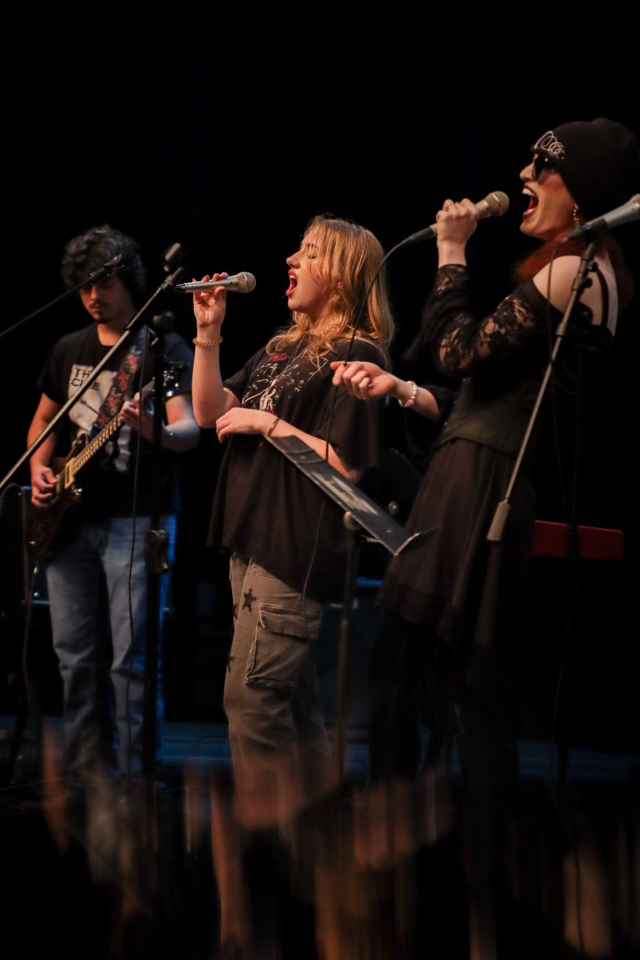 A band performing on stage, featuring a guitarist and two vocalists passionately singing into microphones.