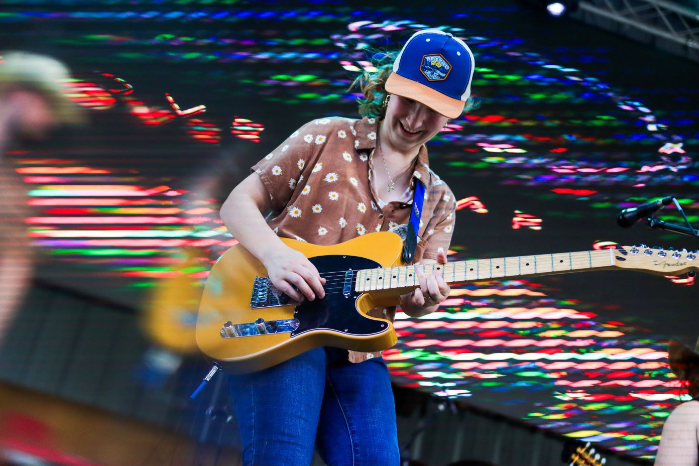 A musician joyfully playing an electric guitar on stage, wearing a floral shirt and a colorful cap.