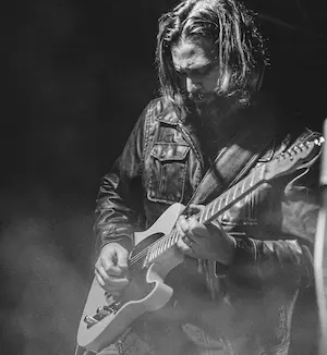 Black-and-white photo of a long-haired guitarist in a leather jacket playing an electric guitar amid stage smoke.