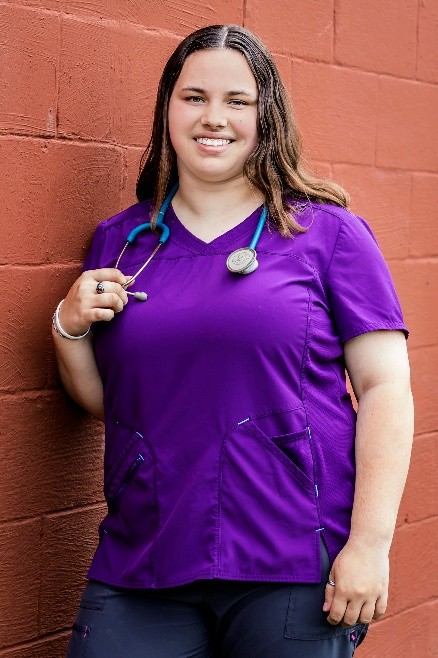 A smiling healthcare professional wearing a purple scrubs top with a stethoscope around her neck, posed against a red wall.