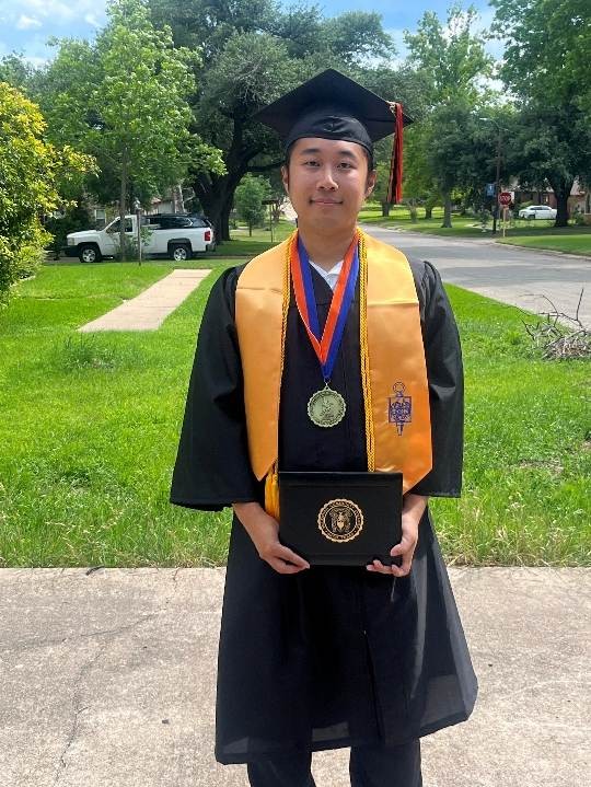 A graduate wearing a black cap and gown with a yellow stole and medals, holding a diploma in front of a grassy area and street.