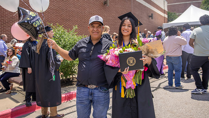 A female graduate stands next to her family member smiling and holding balloons and flowers.