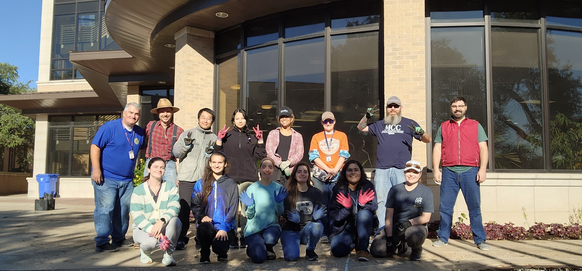 Students, faculty, and staff wearing gardening gloves in front of a building with flower beds.