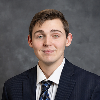 Head-and-shoulders professional portrait of a young man in a dark suit and patterned tie against a gray background.