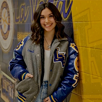 Smiling student wearing a blue-and-gray letterman jacket, standing against a yellow school hallway wall.