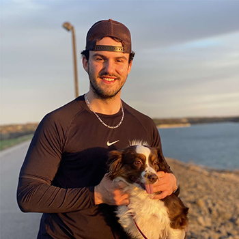 Smiling man in a black athletic shirt and backward cap holding a brown-and-white dog by the water at sunset.