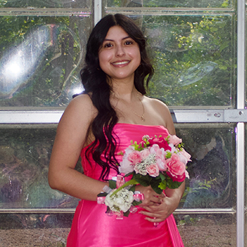 Smiling young woman in a bright pink dress holding a bouquet of pink and white flowers in front of a window.