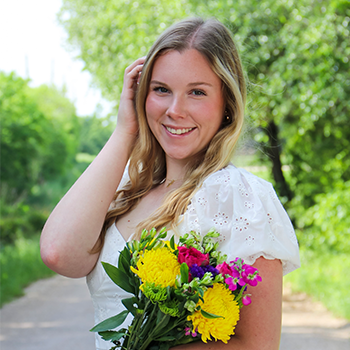 Smiling woman in a white dress holding a colorful bouquet on a tree-lined path.