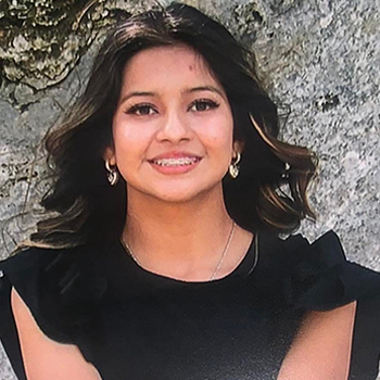 A young woman with long, wavy hair and earrings smiles at the camera while wearing a black top against a textured stone background.