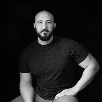 Black-and-white portrait of a bald, bearded man in a dark T-shirt seated against a dark background.