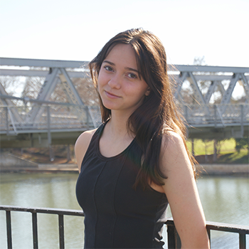 Woman with long dark hair in a black sleeveless top standing by a riverside railing, with a metal bridge in the background.