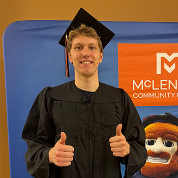 Graduate in cap and gown giving two thumbs up in front of a McLennan Community College banner.