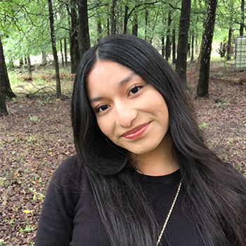 Smiling woman with long dark hair standing in a wooded area.