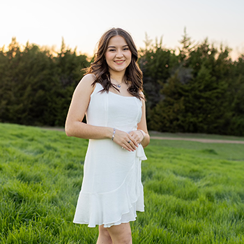 Young woman in a white dress standing in a grassy field at sunset with trees in the background.