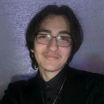 A young man in a black dress shirt and glasses smiles at the camera against a textured background.