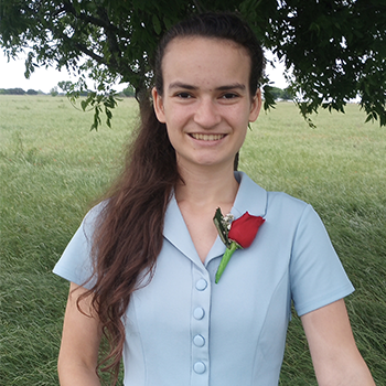 Smiling person standing outdoors in a grassy field under a tree, wearing a light blue shirt with a red rose pinned on the chest.