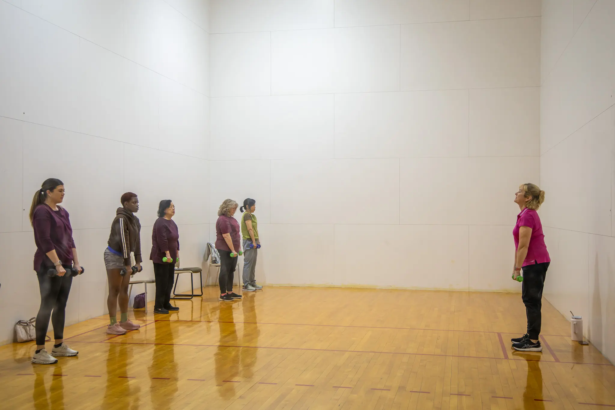A group of women stands in a line, facing a fitness instructor, in a spacious workout area. Each woman holds a dumbbell, indicating a strength training session.