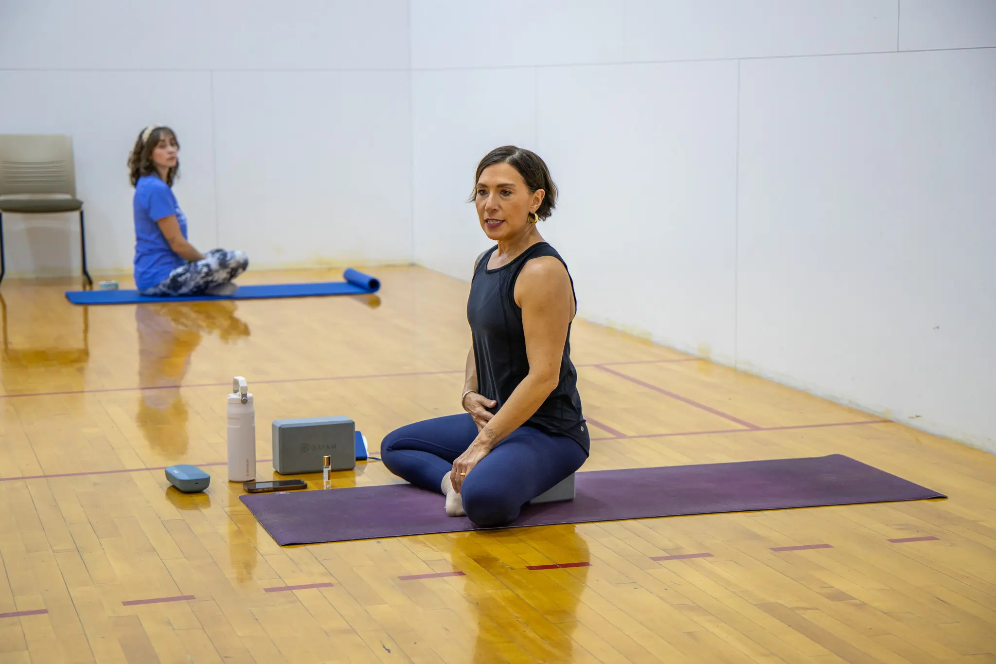 Two women practicing yoga in a gym setting, one sitting on a mat while the other is in the background.